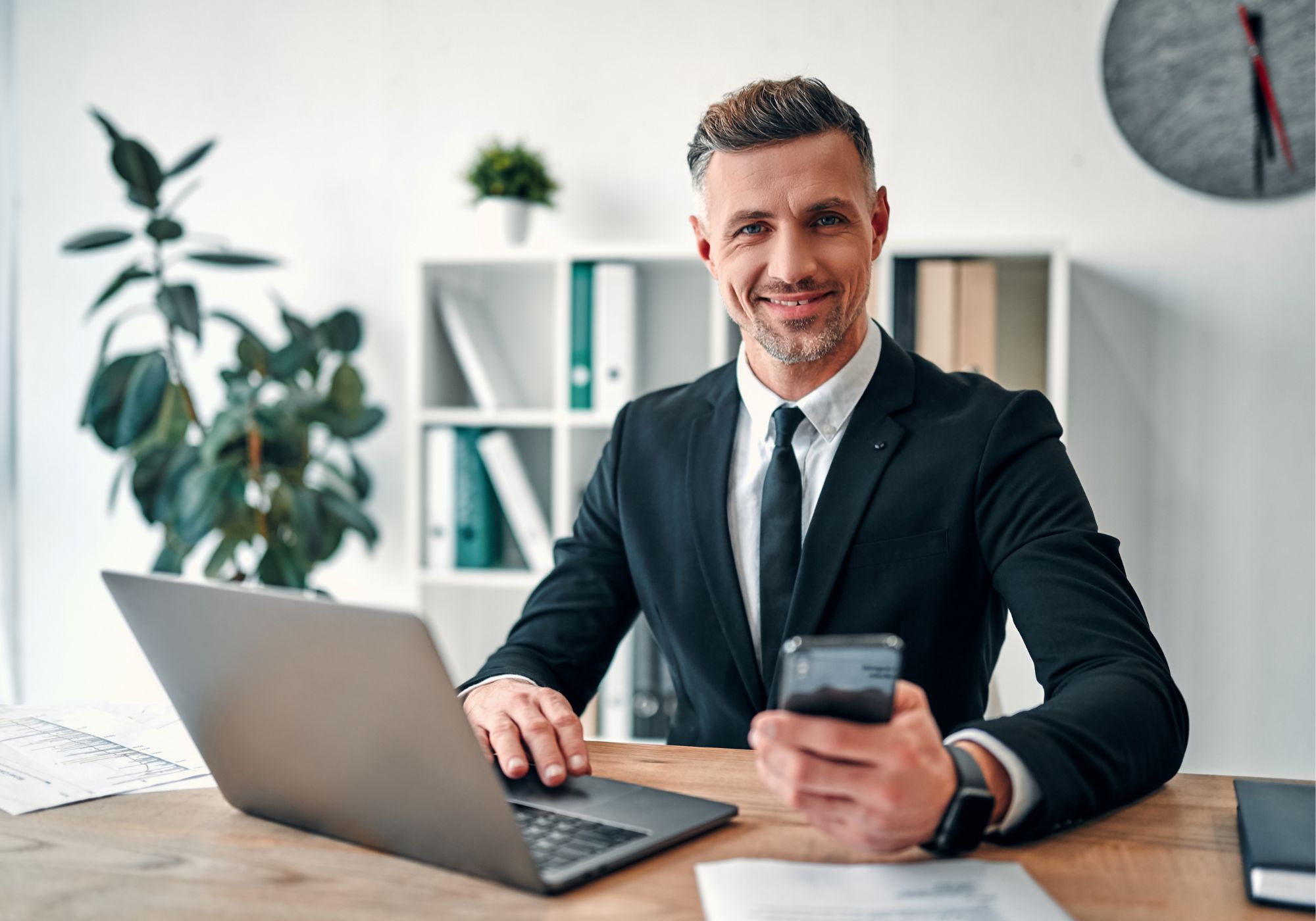 A professional-looking man in a suit sits at a desk with a laptop and papers, holding a smartphone and smiling in a modern office setting.