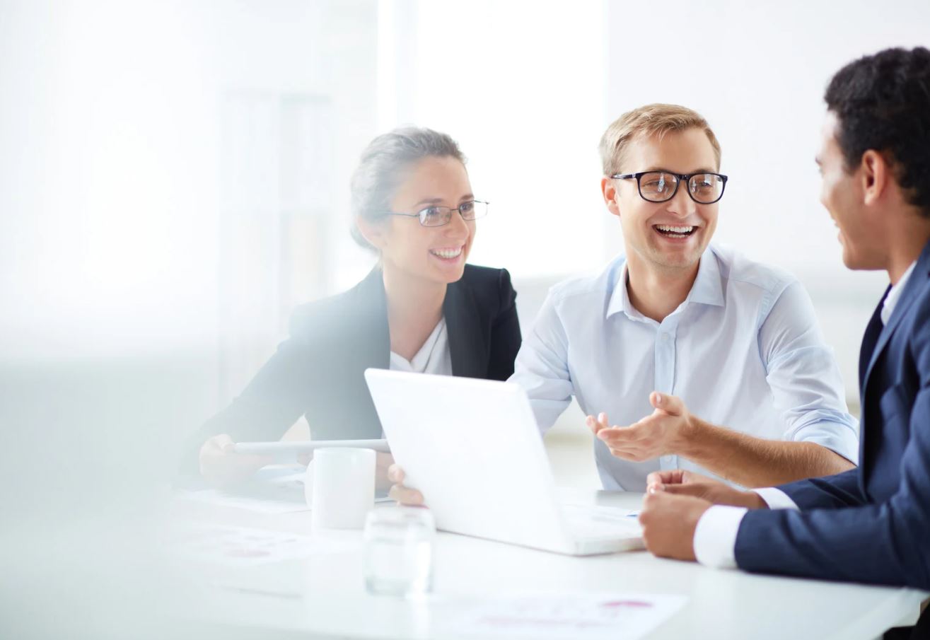 A group of professionals is engaged in a friendly discussion around a laptop in a bright office setting.