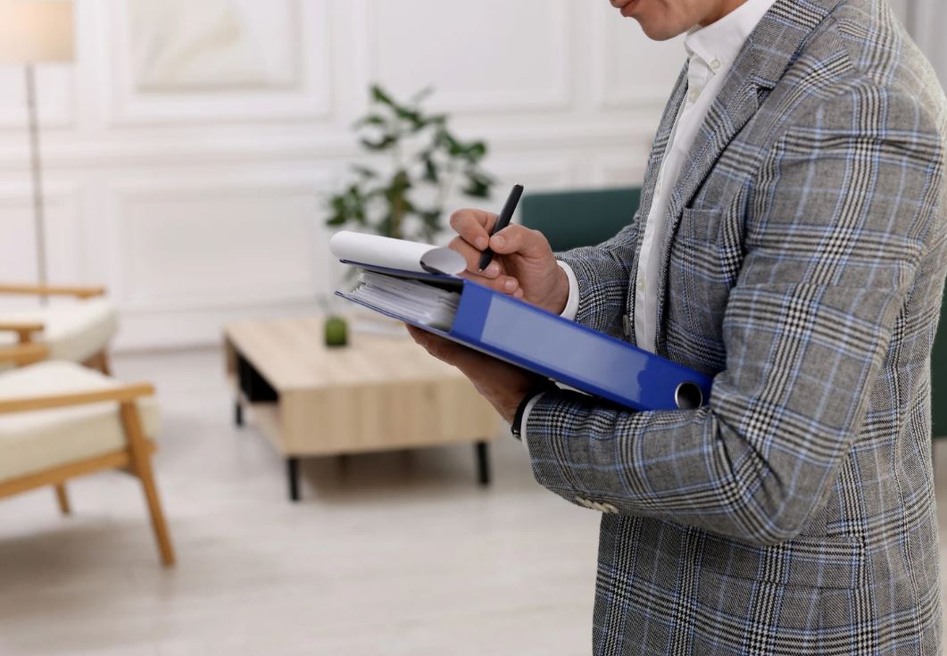 A professional man in a plaid suit holds a blue binder while taking notes with a pen, standing in a modern office setting. The background features light-colored walls, a green plant, and a wooden coffee table, creating a sophisticated workspace atmosphere. This image conveys themes of organization, productivity, and professionalism, ideal for content related to business, office management, or productivity tips.